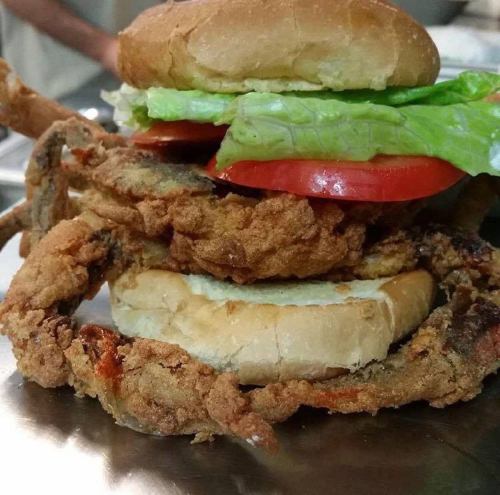A stacked sandwich featuring a fried crab, lettuce, tomato, and a bun on top, served on a metal plate.