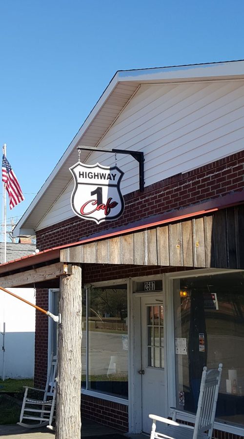 Sign for "Highway 1 Cafe" on a brick building, with an American flag in the background. Rocking chairs on the porch.