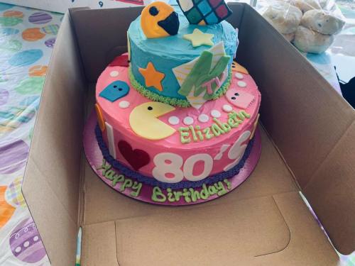 A colorful two-tier birthday cake with playful decorations, including Pac-Man, stars, and the name "Elizabeth."