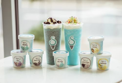 Two colorful drinks topped with whipped cream and sprinkles, surrounded by various ice cream tubs on a table.