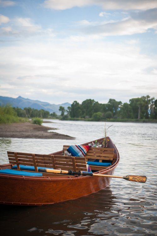 A wooden rowboat with colorful blankets rests on calm water, surrounded by trees and mountains under a cloudy sky.