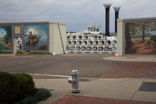 A riverboat is framed by murals on walls, with a fire hydrant in the foreground and a cloudy sky above.