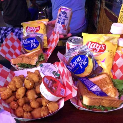A table with two baskets of food: fried snacks, sandwiches, chips, and Moon Pies, alongside cans of beer.