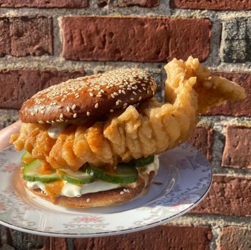 A whimsical sandwich featuring a fried fish shaped like a whale, layered with pickles and sauce on a sesame seed bun.