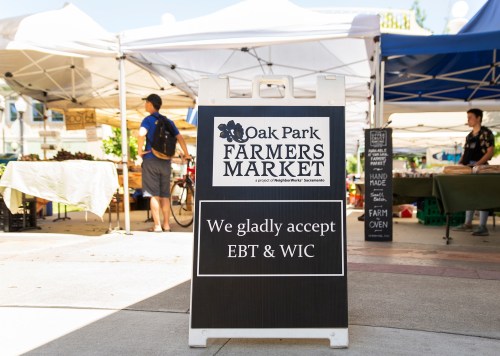 Sign for Oak Park Farmers Market, stating they accept EBT and WIC, with market stalls and people in the background.