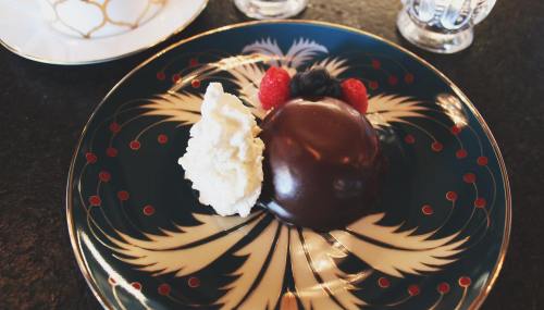 A chocolate dessert dome on a decorative plate, garnished with whipped cream, raspberries, and blueberries.