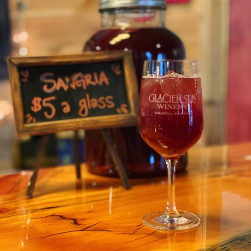 A glass of sangria on a wooden table, with a sign advertising it for $5 at Glacier Sun Winery.