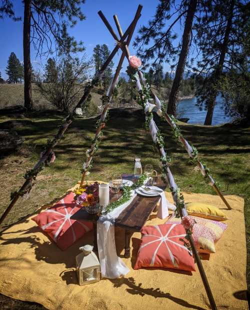 A cozy outdoor picnic setup with a wooden table, colorful pillows, and floral decorations near a river.