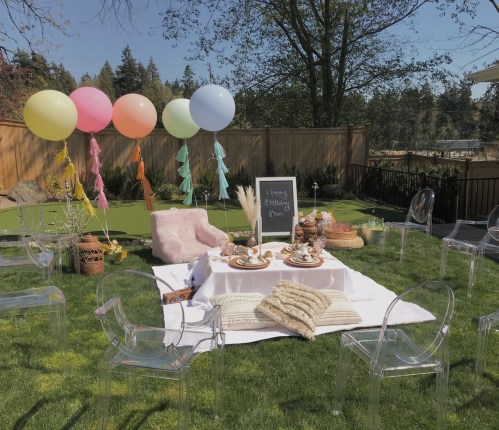 A cozy outdoor picnic setup with balloons, a table, snacks, and decorative elements on a grassy area.