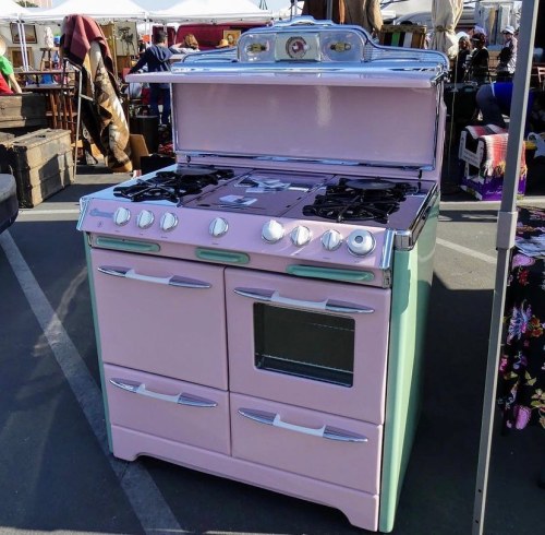 A vintage pink and green gas stove with multiple burners and an oven, displayed at an outdoor market.