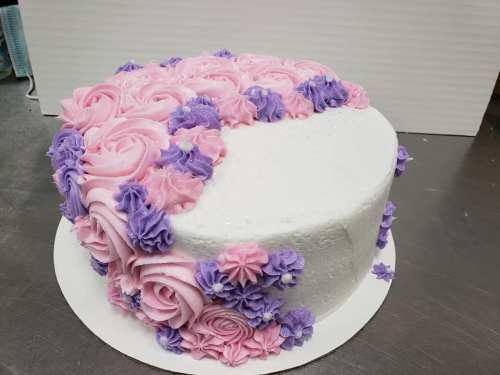 A round cake decorated with pink and purple buttercream roses and flowers on one side, with a smooth white finish.