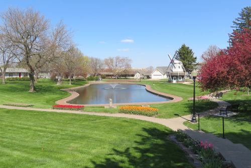 A serene park scene featuring a pond, colorful flower beds, and trees under a clear blue sky.