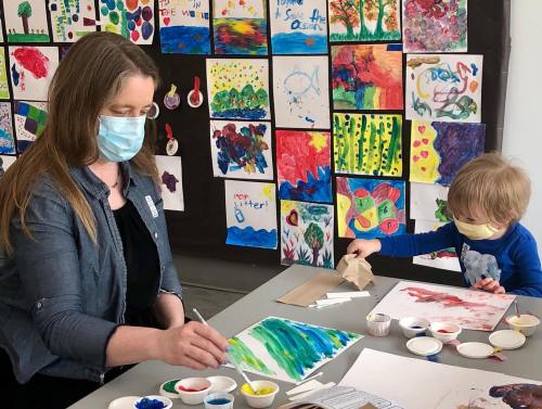 A woman and a child paint together at a table, surrounded by colorful artwork on the wall. Both are wearing masks.