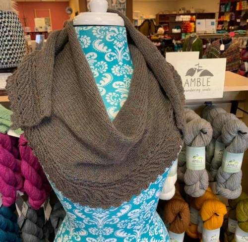 A knitted shawl displayed on a mannequin, surrounded by colorful yarns in a cozy craft shop.
