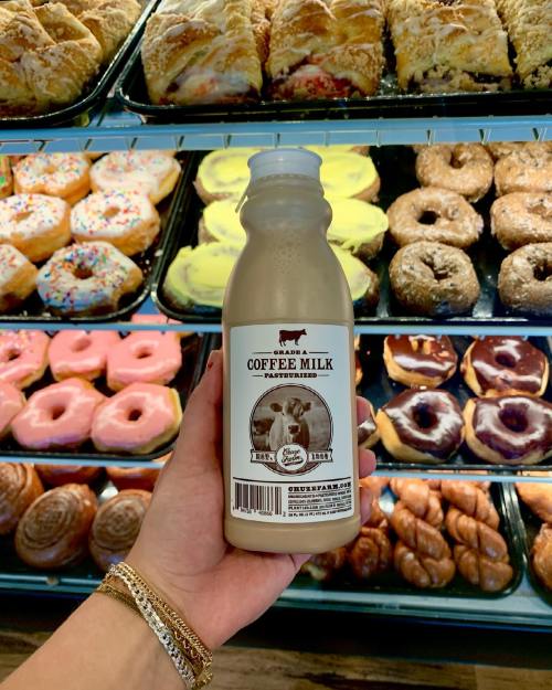 A hand holds a bottle of coffee milk in front of a display of colorful donuts and pastries.