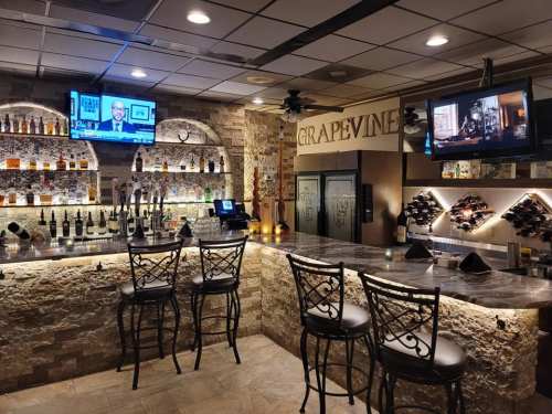 A cozy bar area with stone walls, a marble countertop, and two TVs displaying news and a movie.