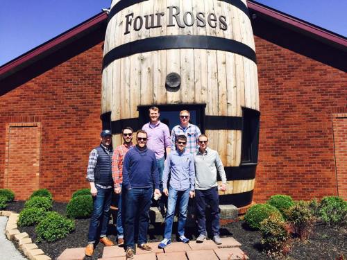 A group of seven men poses in front of a large wooden barrel at the Four Roses distillery on a sunny day.