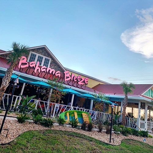 Exterior of a colorful restaurant named "Bahama Breeze," featuring palm trees and outdoor seating under blue skies.