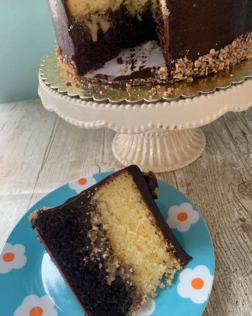 A slice of layered chocolate and vanilla cake on a blue plate with flowers, next to a whole cake on a stand.