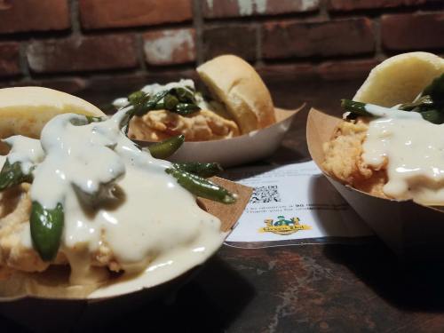 Three fried chicken sandwiches topped with green beans and creamy sauce, served in paper trays on a dark surface.