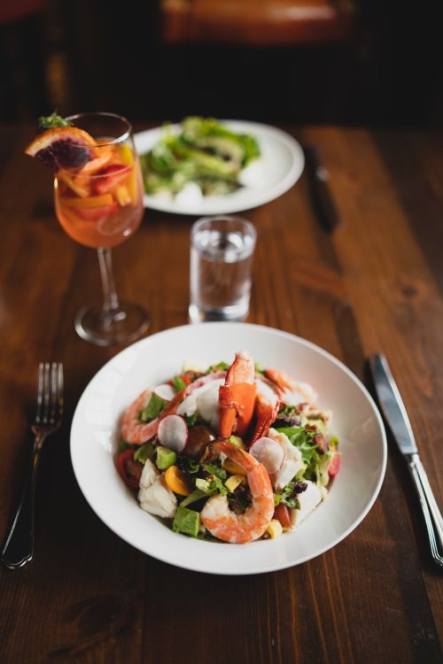 A vibrant salad with shrimp, mixed greens, and colorful vegetables, accompanied by a drink and a second plate in the background.