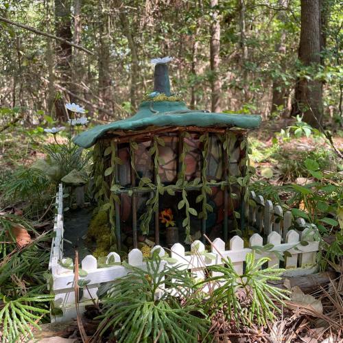 A whimsical miniature house surrounded by greenery and a white picket fence in a forest setting.