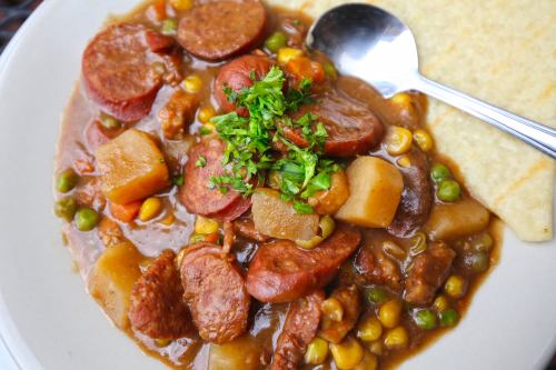 A hearty dish featuring sausage, potatoes, corn, and peas, garnished with fresh herbs, served with a side of flatbread.