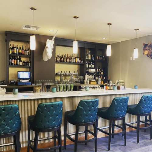 A stylish bar with a white unicorn head on the wall, featuring green upholstered chairs and a variety of liquor bottles.