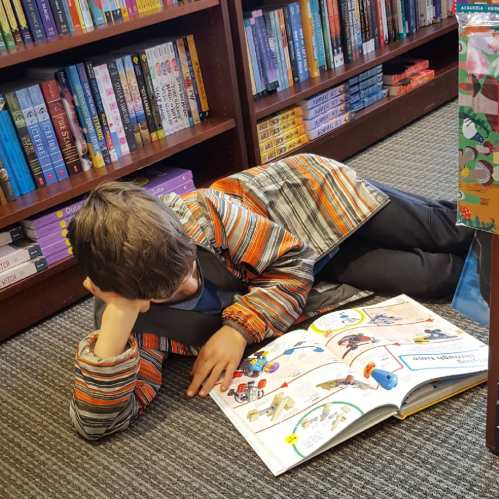 A child in a striped jacket lies on the floor, reading a colorful book in a cozy bookstore.