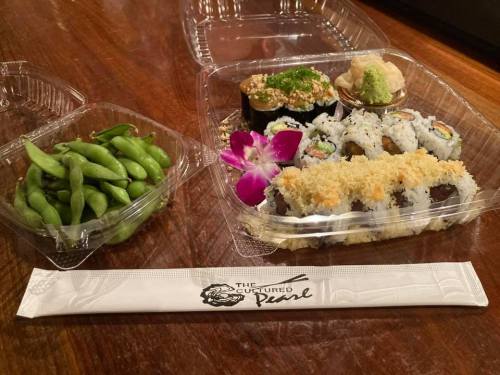 A takeout sushi platter with rolls, edamame, and a flower, accompanied by a chopstick wrapper from The Cultured Pearl.