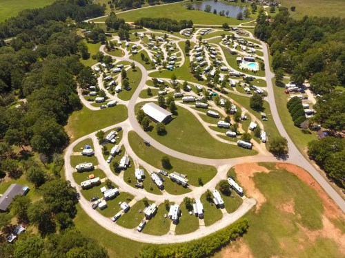 Aerial view of a large RV park with winding roads, grassy areas, and a pond surrounded by trees.