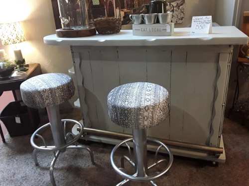 A rustic bar with a white top and two patterned stools, featuring a sign that says "Eat Local" priced at $550.
