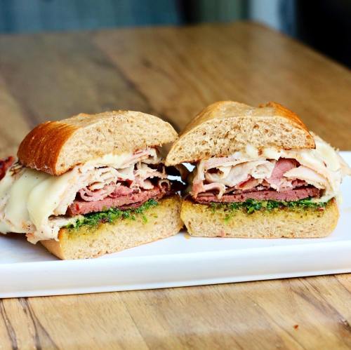 A close-up of a sandwich with layers of turkey, roast beef, and cheese on whole grain bread, served on a white plate.