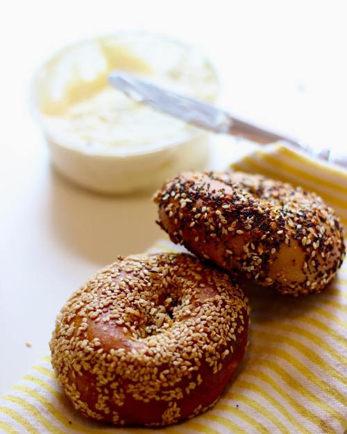 Two sesame bagels on a striped napkin, with a bowl of cream cheese and a knife in the background.