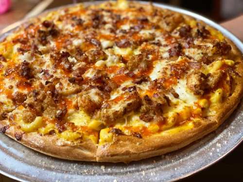 A close-up of a pizza topped with cheese, crispy chicken, bacon, and a drizzle of sauce on a golden crust.
