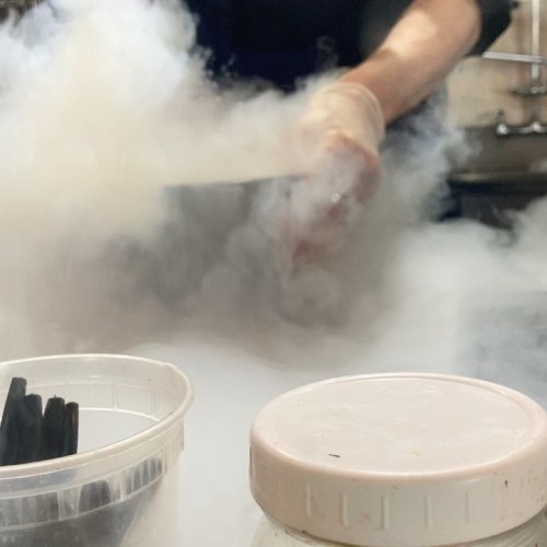 A hand in gloves is seen amidst swirling smoke, with containers in the foreground, suggesting a cooking or scientific process.