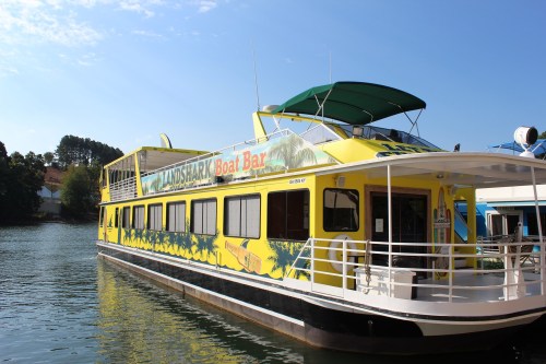 A bright yellow boat bar named "Landshark" docked on calm water, surrounded by trees and blue sky.
