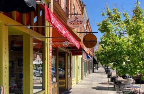 A sunny street view featuring storefronts, including a bakery and a bookstore, with trees lining the sidewalk.