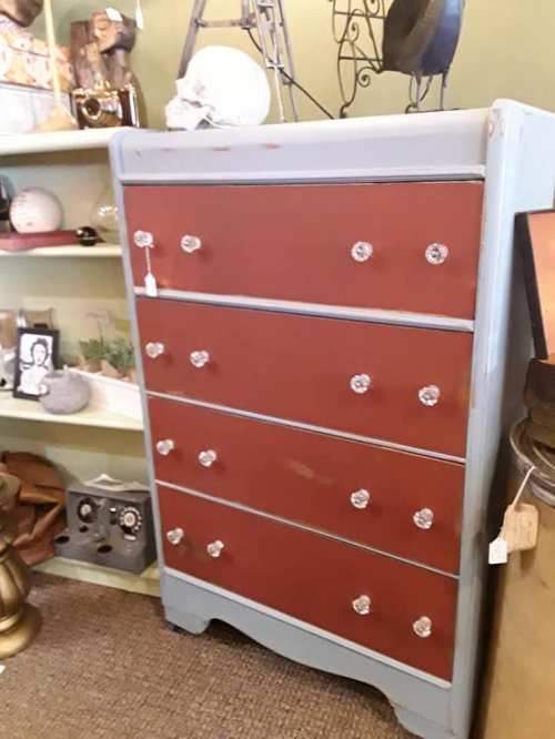 A vintage dresser with four red drawers and clear crystal knobs, set against a colorful display of decor items.