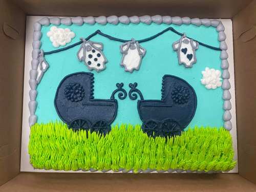 A decorated cake featuring two baby carriages, hanging onesies, and a blue sky with clouds, set on green grass.