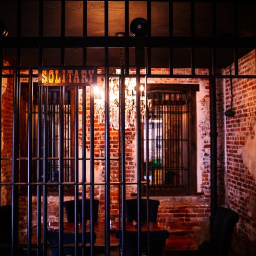 A dimly lit room with exposed brick walls, featuring a barred entrance labeled "SOLITARY" and a chandelier above.