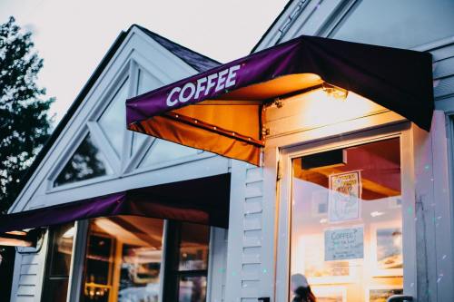 A cozy coffee shop entrance with a purple awning and warm lighting, surrounded by a charming exterior.