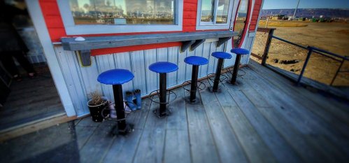 Five blue bar stools lined up against a red and white building, with a wooden deck and a scenic view in the background.