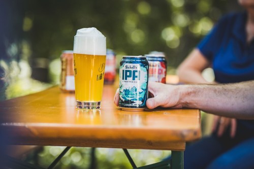 A glass of beer and a can of IPF beer on a wooden table, with blurred background and other cans visible.