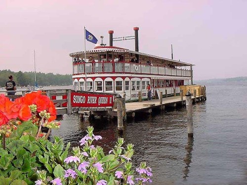 A riverboat docked at a pier, surrounded by flowers, with a flag flying and calm waters in the background.