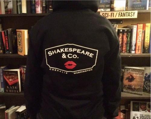 A person wearing a black hoodie with "Shakespeare & Co." and a red lip logo, standing in front of bookshelves.