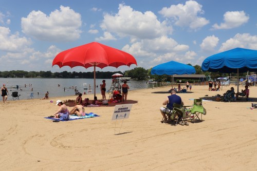 A sandy beach with colorful umbrellas, people relaxing, and a lake under a blue sky with fluffy clouds.