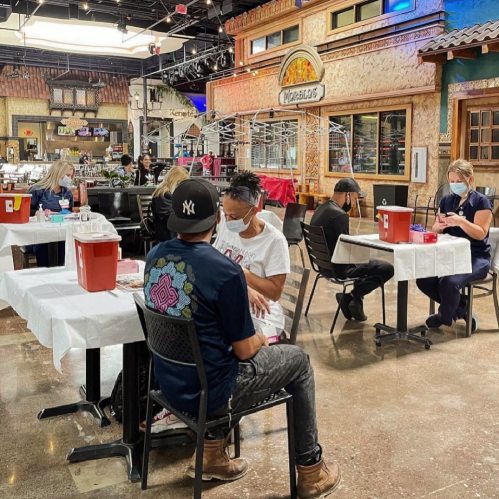 People seated at tables in a community space, participating in a vaccination event, wearing masks and social distancing.