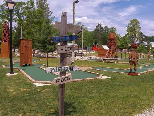 A signpost points to restrooms, rides, and mini-golf in a vibrant outdoor amusement area with greenery and attractions.