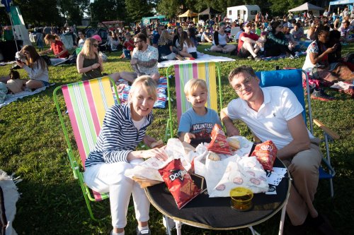 A family enjoys snacks on a picnic blanket at a crowded outdoor event, smiling and sitting in colorful chairs.